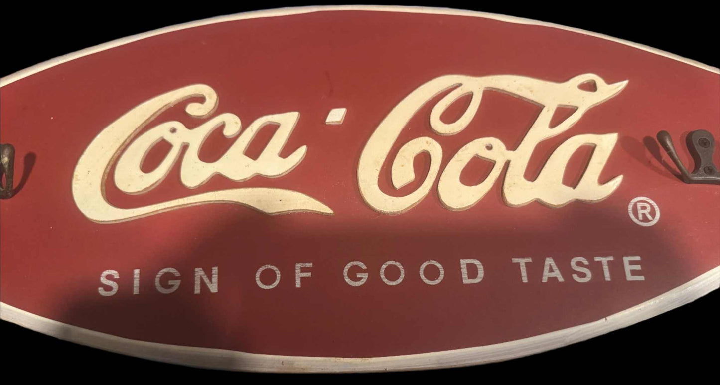 Coca-Cola wooden wall plaque with "Sign of Good Taste" text and hooks for hanging keys or accessories, vintage style 19x8 inches.